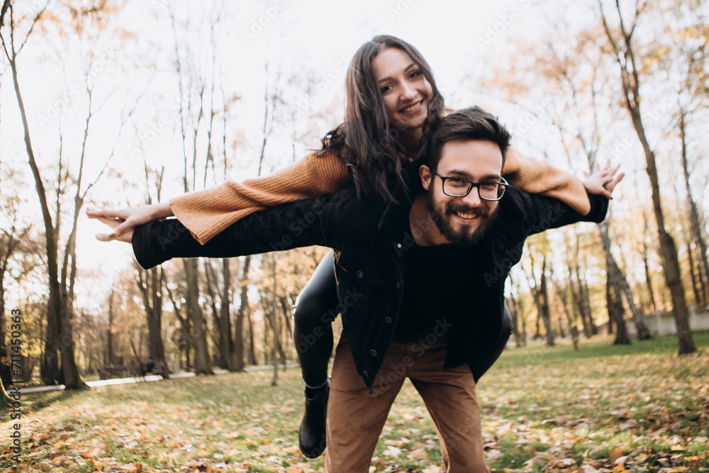Heterosexual caucasian young loving couple, husband carrying on back his wife hands wings fly, hugging smiling kissing laughing spending time together. Autumn, fall season, orange yellow red maple lea - obrazy, fototapety, plakaty 