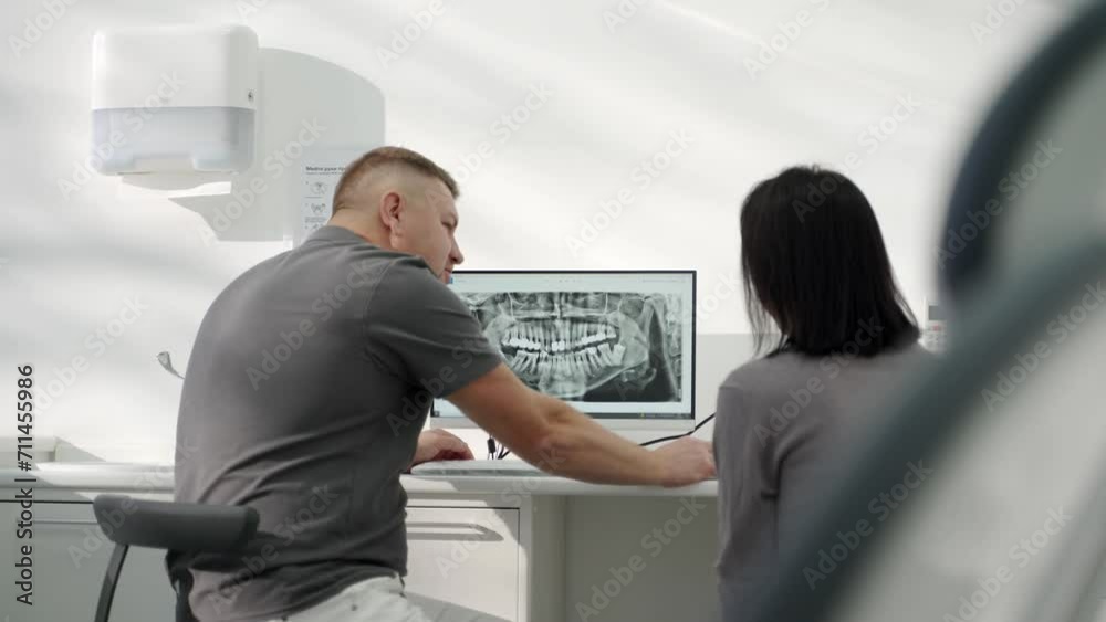 Male dentist showing x-ray photo of jaws on computer screen to female patient in dental clinic. Orthodontist explaining x-ray of teeth on screen to woman. Consultation with dental surgeon. Back view. Male dentist showing x-ray photo of jaws on computer screen to female patient in dental clinic. Orthodontist explaining x-ray of teeth on screen to woman. Consultation with dental surgeon. Back view.