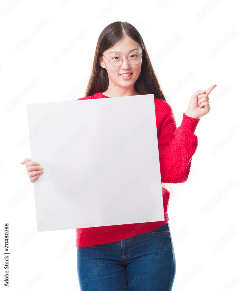 Young Chinese woman over isolated background holding blank banner very happy pointing with hand and finger to the side Young Chinese woman over isolated background holding blank banner very happy pointing with hand and finger to the side
