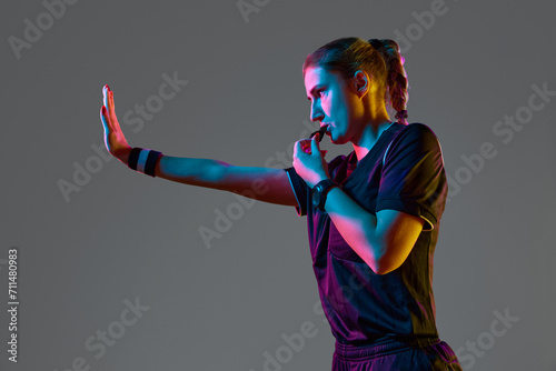 Athletic pose, young woman referee blowing whistle with serious expression against grey studio background in neon light. Concept of sport, competition, match, profession, football game, control photo
