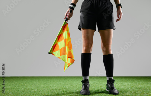 Cropped image of female legs, referee on field standing with flag to signal game rules against grey studio background. Concept of sport, competition, match, profession, football game, control photo