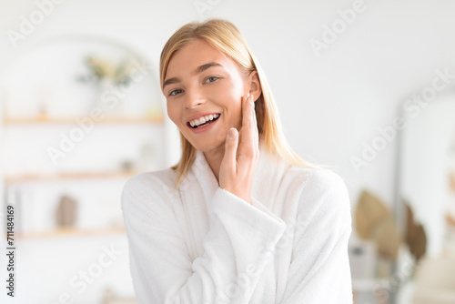 Young blonde woman touches smooth cheek after skincare ritual indoors