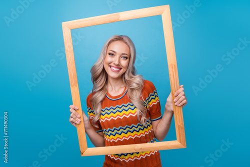 Photo of adorable pretty girl dressed print shirt holding wooden frame isolated blue color background