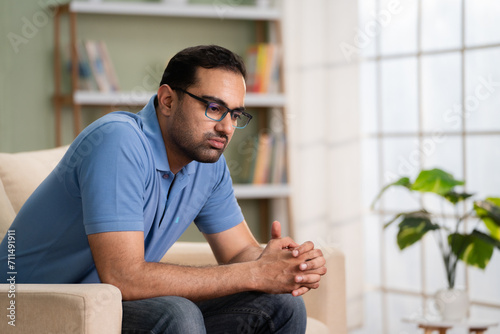 Worried indian man in deep thinking while sitting on sofa at home - concept of financial problem, love breakup and mental illness.
