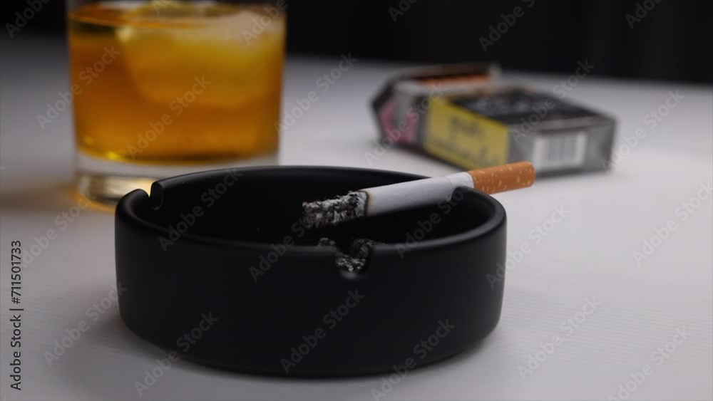 Time lapse of burning cigarette in a black ashtray and a whiskey glass in the background Time lapse of burning cigarette in a black ashtray and a whiskey glass in the background