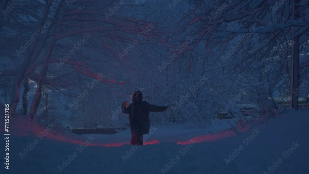 A millennial girl holds a red burning signal light in her hand. A woman in a snowy winter forest, among the trees in a national park or nature reserve. Fire or flame. The female enjoys the snowfall.