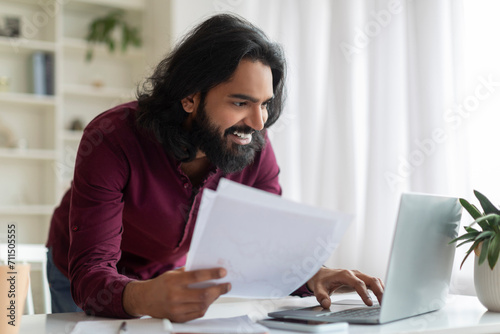 Focused indian man analyzing documents and working on his laptop at home