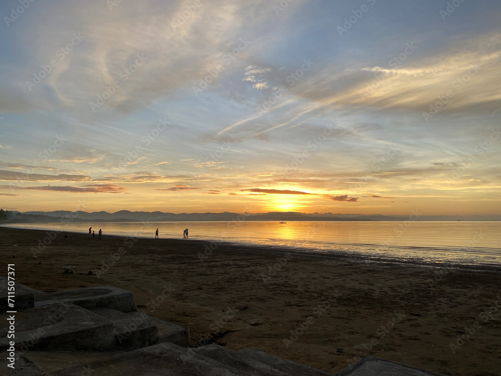 Obraz premium On the beach, the golden early sunrise casts clouds on the sea horizon. Unrecognizable figures enjoy the view. Silhouettes