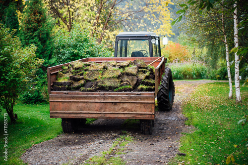 Removed soil, lawn, turf in a tractor trailer in a dense green garden. Transportation and replacement of land on lawns. Gardening and agricultural machinery