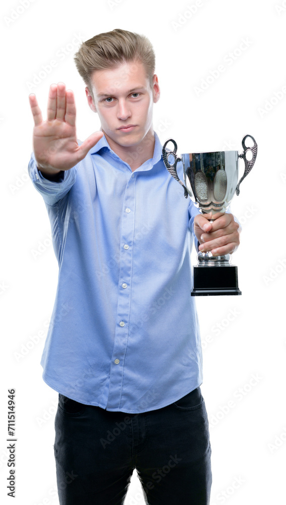 Young handsome blond man holding a trophy with open hand doing stop sign with serious and confident expression, defense gesture