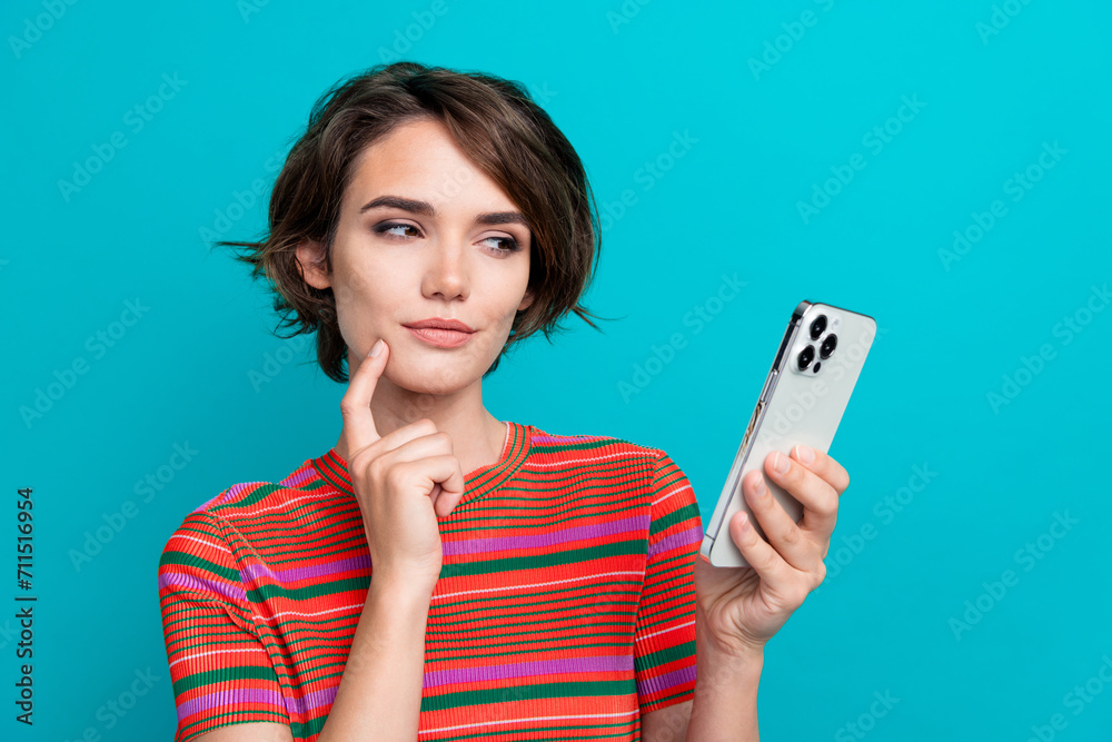 Photo of minded intelligent woman dressed striped t-shirt look at smartphone finger on cheek isolated on turquoise color background Photo of minded intelligent woman dressed striped t-shirt look at smartphone finger on cheek isolated on turquoise color background