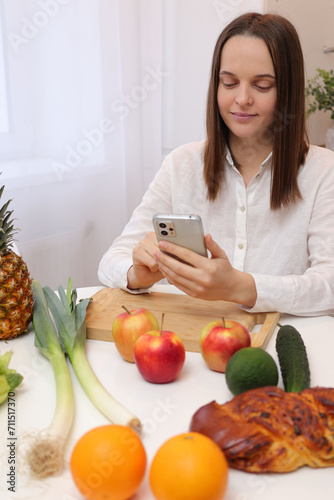 Charming attractive brown haired woman in white shirt sitting at table with fruits and vegetables scrolling online via smartphone using mobile app with healthy nutrition checklist