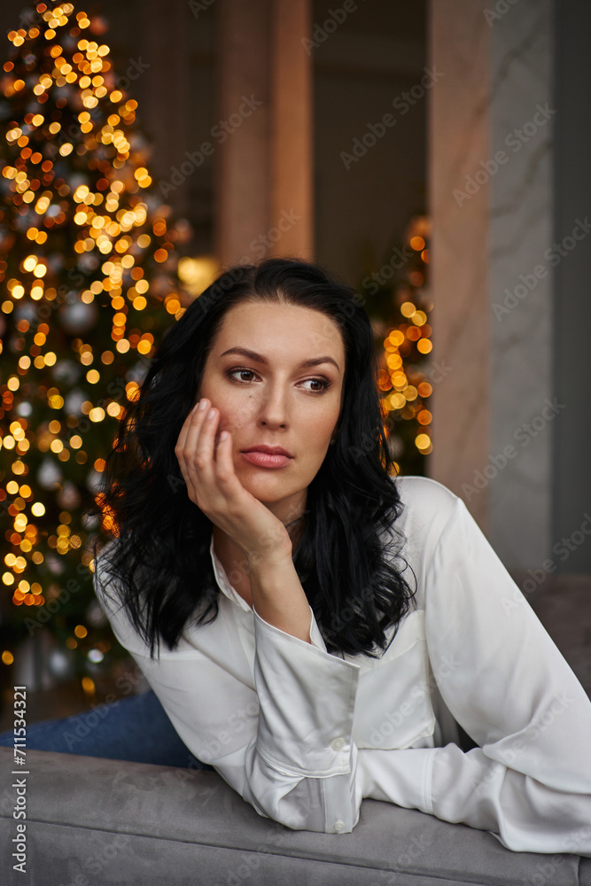 Fototapeta premium Gracefully seated in a grey chair, the young woman in a white shirt captivates against the backdrop of sparkling lights from a Christmas tree.