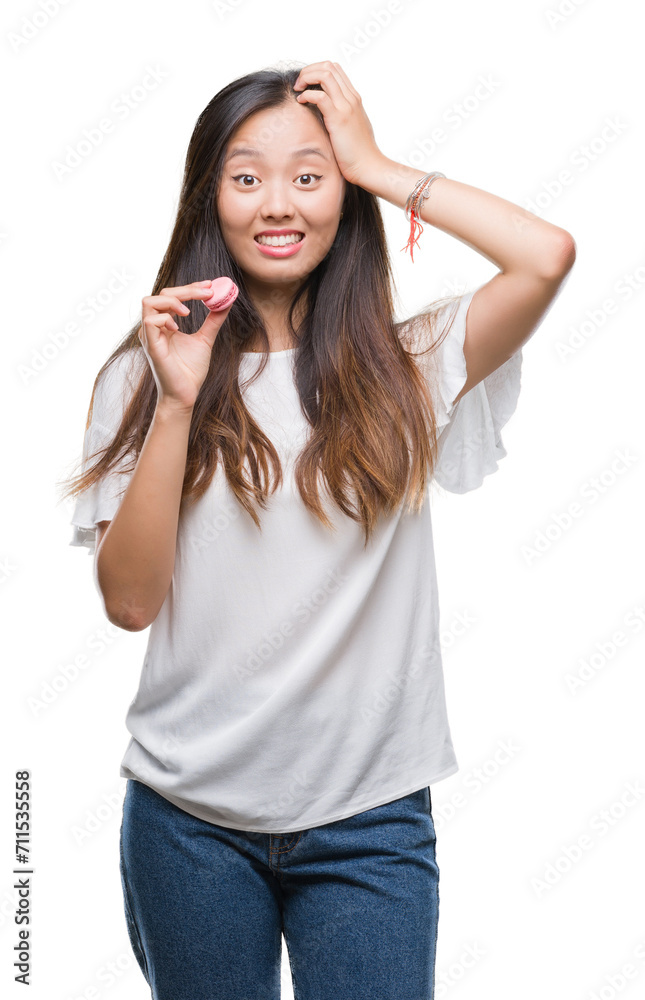 Young asian woman eating pink macaron sweet over isolated background stressed with hand on head, shocked with shame and surprise face, angry and frustrated. Fear and upset for mistake. Young asian woman eating pink macaron sweet over isolated background stressed with hand on head, shocked with shame and surprise face, angry and frustrated. Fear and upset for mistake.