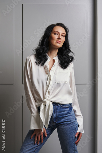  Set in a stylish interior with a grey wall, the dark-haired beauty in a white shirt and jeans captures attention, defining the contemporary aesthetic.