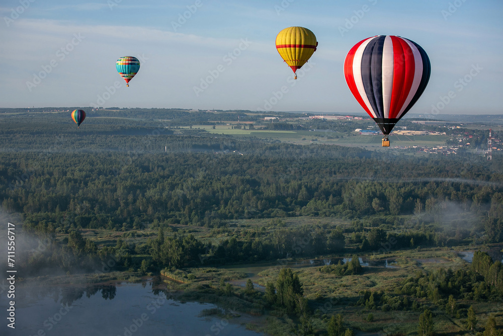 Colorful hot air balloons fly over the river and forest. A small town is visible in the distance. Early summer morning. Balloon flight.