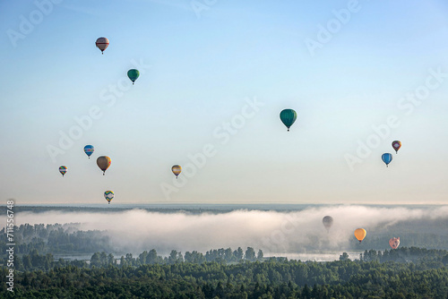 Lots of colorful hot air balloons against the blue sky. Below the balls is a forested area and a river covered in fog. Early summer morning. Balloon flight.