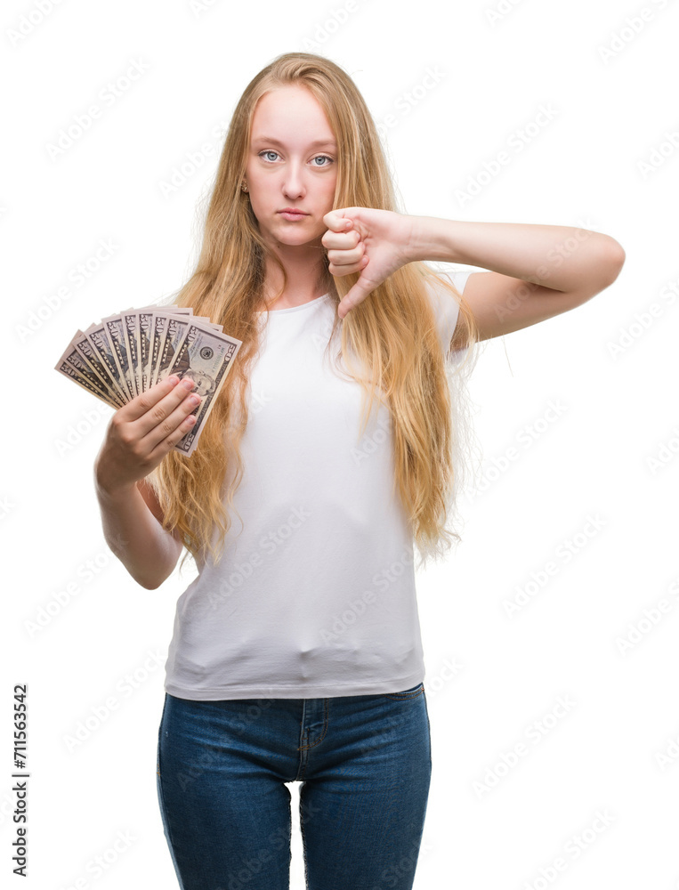 Blonde teenager woman holding dollars with angry face, negative sign showing dislike with thumbs down, rejection concept Blonde teenager woman holding dollars with angry face, negative sign showing dislike with thumbs down, rejection concept