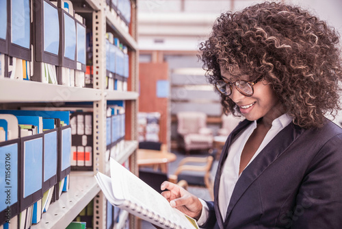 Young African corporate woman looking through shelves with binders and books in large start-up office space. Cape Town, South Africa