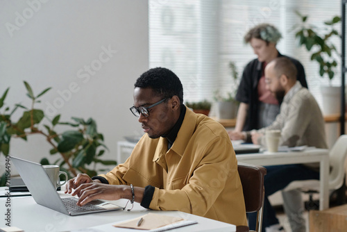 Black man in casual outfit typing on laptop at office desk, his colleagues working on background