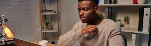 young concentrated african american freelancer working on laptop in evening at home, banner photo