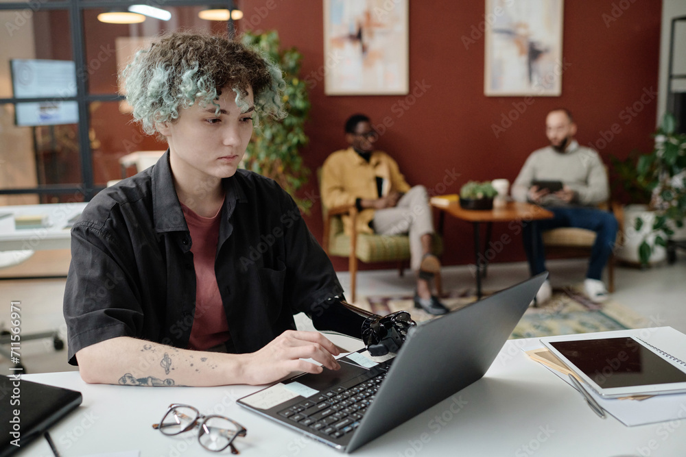 Young girl with prosthetic arm browsing laptop at office, her male colleagues sitting on background Young girl with prosthetic arm browsing laptop at office, her male colleagues sitting on background