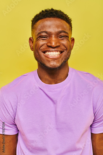 jolly african american man in purple t-shirt with radiant smile looking at camera on yellow backdrop