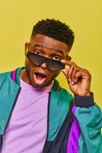 impressed african american man with open mouth holding sunglasses and looking at camera on yellow photo