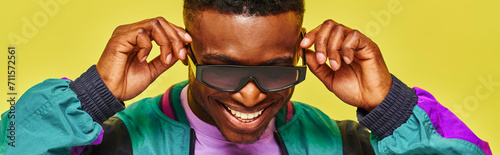 trendy african guy in bright jacket adjusting sunglasses and smiling on yellow backdrop, banner photo