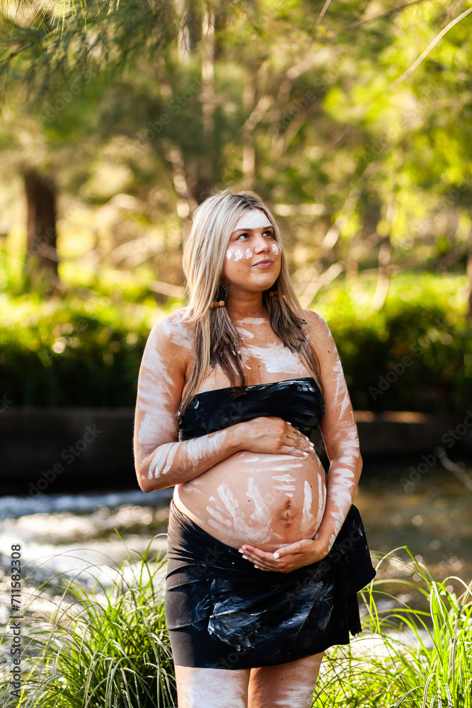 Pregnant Aboriginal lady in ochre body paint standing by water in bushland looking upwards Pregnant Aboriginal lady in ochre body paint standing by water in bushland looking upwards