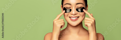 Skin care eye patches. Asian beauty care concept. Beautiful model applying black hydro gel collagen mask on her eyes and smiling against green background