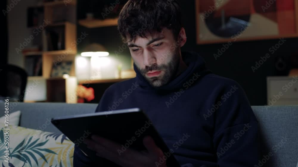 Bearded male sitting at sofa and holding tablet. Handsome man using social media. Male holding device and browsing. Man conducting online purchase. Male searching in Internet.