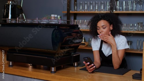 A dark-skinned young woman is bored at the counter of a coffee shop, scrolling her feed on social networks, sitting on the phone yawning yawning dark-skinned woman she is bored there are no customers.