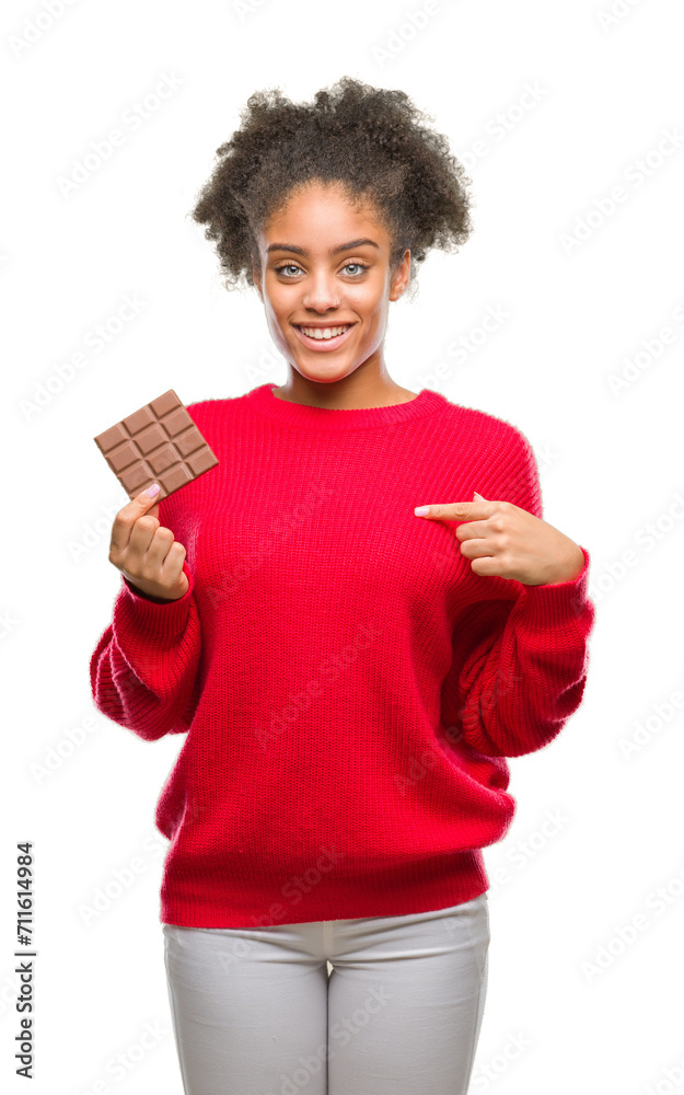 Young afro american woman eating chocolate bar over isolated background with surprise face pointing finger to himself Young afro american woman eating chocolate bar over isolated background with surprise face pointing finger to himself