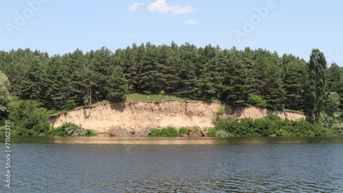 A cliff made of clay near a wide river. A coniferous forest grows above the cliff.