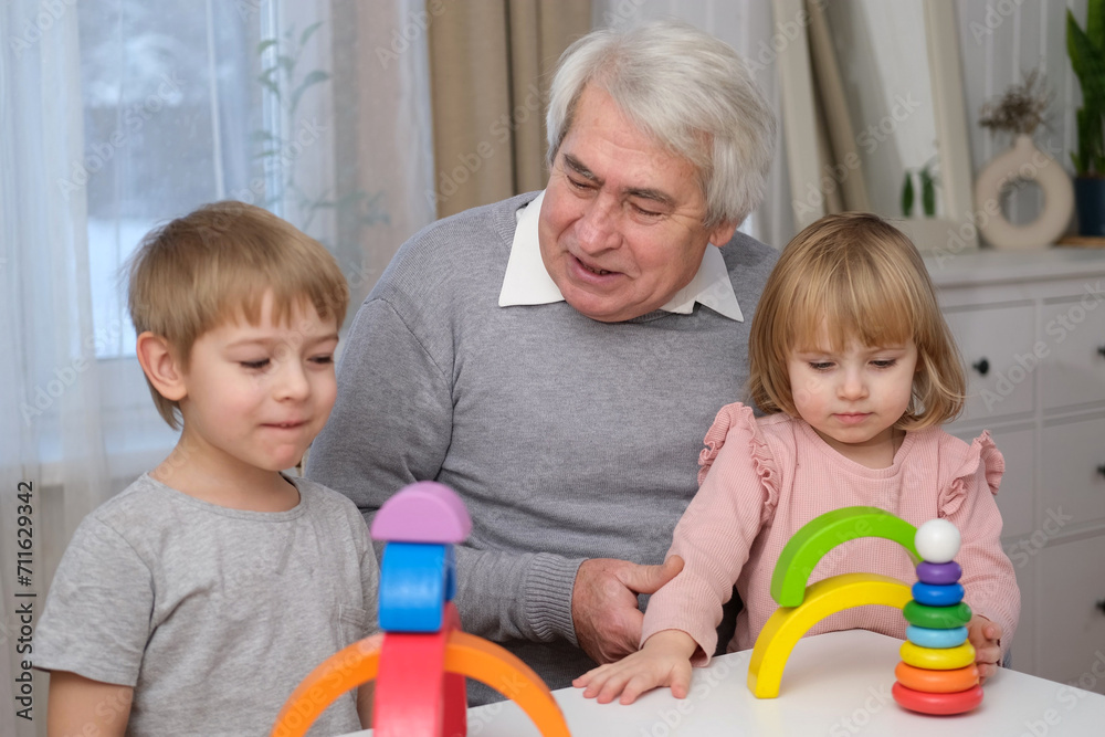Grandfather playing toys with grandson and young granddaughter indoor. Old man grandpa studying with his grandchildren at weekend. Senior male having fun with kids. Happy multigeneration family shot Grandfather playing toys with grandson and young granddaughter indoor. Old man grandpa studying with his grandchildren at weekend. Senior male having fun with kids. Happy multigeneration family shot