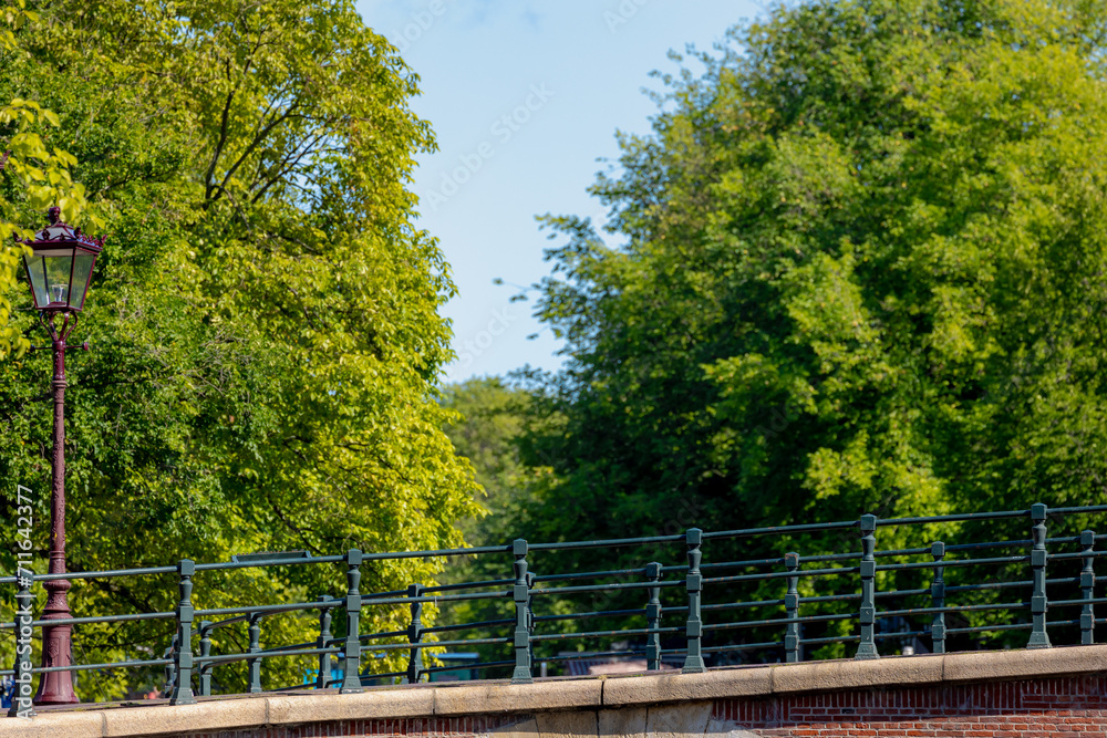 Selective focus of steel railings and brick bridge crossing between the canals, Blurred trees and green leaves along the street under blue clear sky as background, Canals of Amsterdam, Netherlands. Selective focus of steel railings and brick bridge crossing between the canals, Blurred trees and green leaves along the street under blue clear sky as background, Canals of Amsterdam, Netherlands.