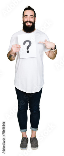 Young man with long hair and bear holding a paper with question mark very happy pointing with hand and finger