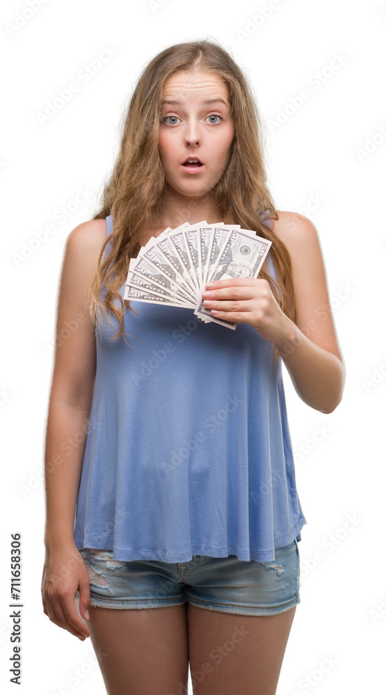 Young blonde woman holding dollars scared in shock with a surprise face, afraid and excited with fear expression Young blonde woman holding dollars scared in shock with a surprise face, afraid and excited with fear expression