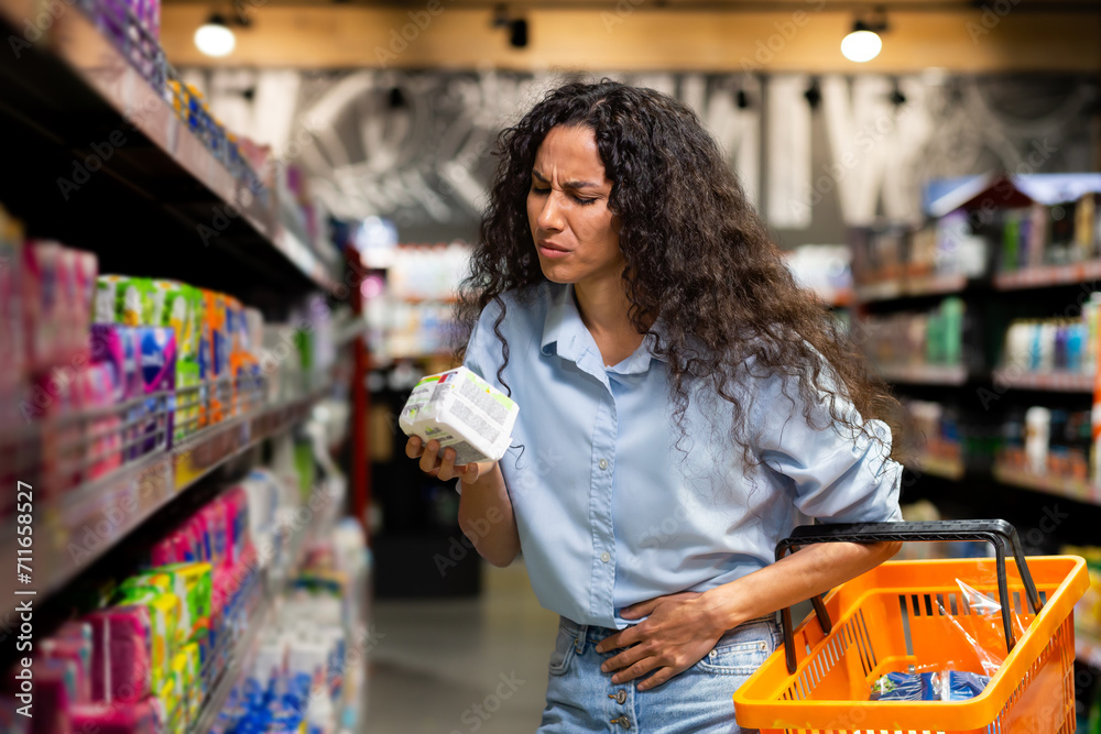 A thoughtful woman with curly hair shops for groceries, analyzing product labels with a pensive look as she holds a basket.