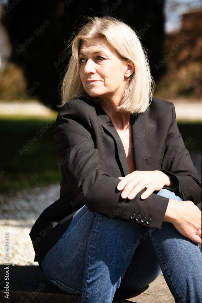 Blonde woman in her mid-fifties portraits in a black jacket, jeans and pumps photographed with the sun aperture wide open in the park. Blonde woman in her mid-fifties portraits in a black jacket, jeans and pumps photographed with the sun aperture wide open in the park.