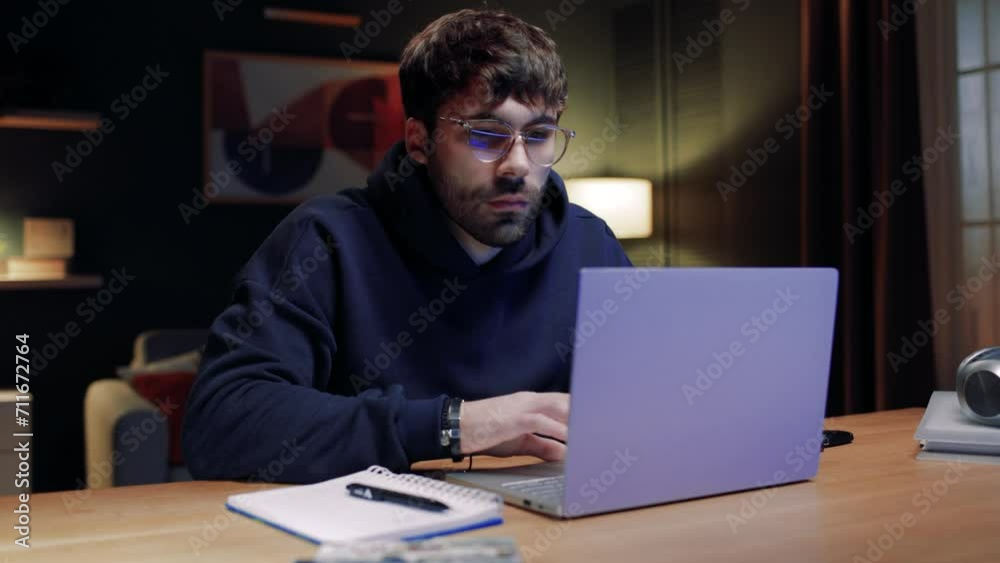 Man sitting at table and using laptop. Bearded male looking at laptop screen. Man satisfied with result of work. Male holding pen and revising finished project. Man spending evening at home.