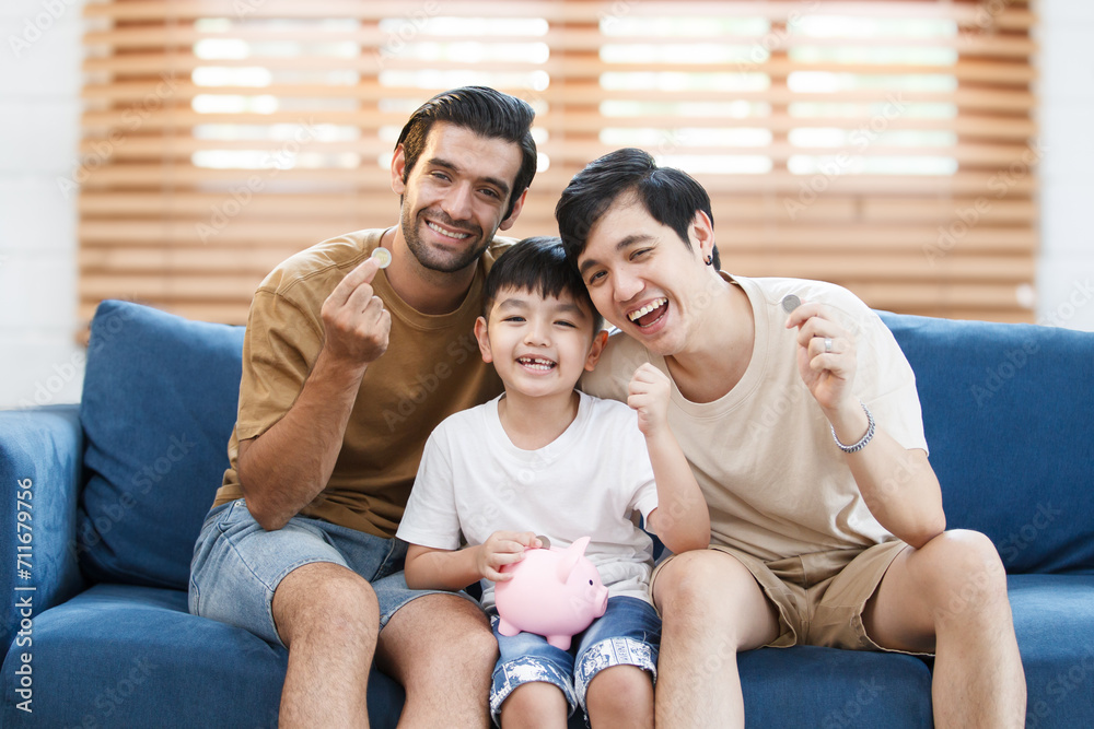 Happy LGBTQ couple with their adopted child in relaxing together in living room, men let the boy learn about saving money and investment. Happy LGBTQ couple with their adopted child in relaxing together in living room, men let the boy learn about saving money and investment.