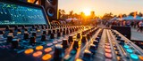 An audio mixing console at a live concert event glows with vibrant controls as the sun sets in the background.
