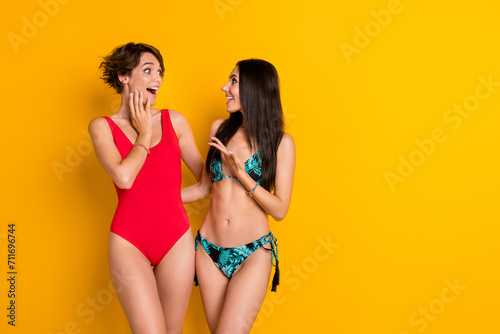 Photo of positive impressed girls dressed swimsuits hugging talking empty space isolated yellow color background