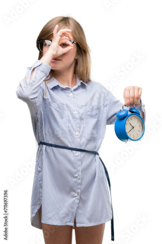 Young blonde child holding alarm clock with happy face smiling doing ok sign with hand on eye looking through fingers