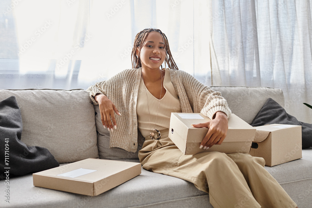 joyful young african american woman in cozy attire sitting on sofa with boxes and smiling at camera joyful young african american woman in cozy attire sitting on sofa with boxes and smiling at camera