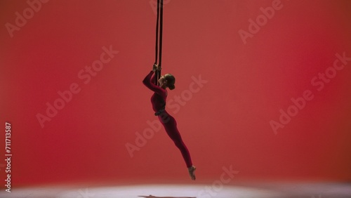 Female gymnast isolated on red studio background. Girl aerial dancer in bodysuit on acrobatic trapeze with straps perform element. photo