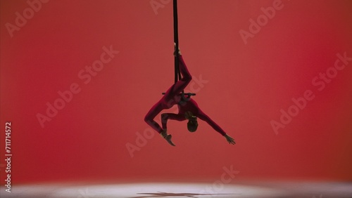 Female gymnast isolated on red studio background. Girl aerial dancer in bodysuit on acrobatic trapeze with straps perform element.