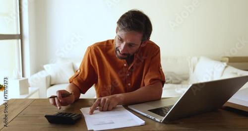 Portuguese middle-aged man doing accountancy work at home, sit at desk calculates expenses, control incomes on calculator, pay household bills, taxes use laptop and e-banking app. Finances management photo